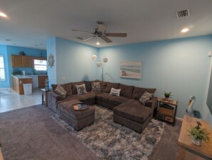 Una sala de estar con un sofá modular, un ventilador de techo, una cocina al fondo y un cuadro con motivos playeros en la pared.
