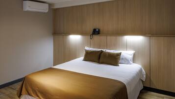 A hotel room with a wooden headboard, a bed with brown and white bedding, a bedside phone, and an air conditioning unit on the wall.