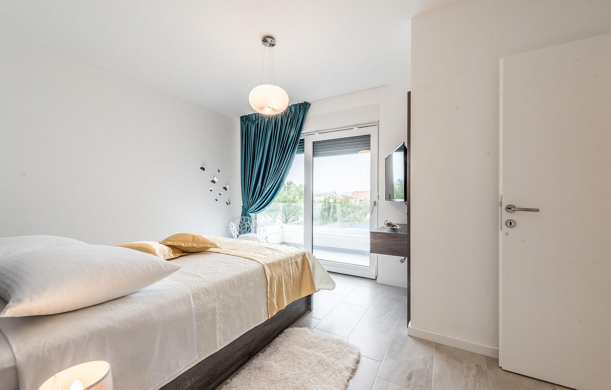 A bedroom with a bed, a nightstand, a mirror, a door, and a view of the outside.