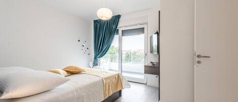 A bedroom with a bed, a nightstand, a mirror, a door, and a view of the outside.