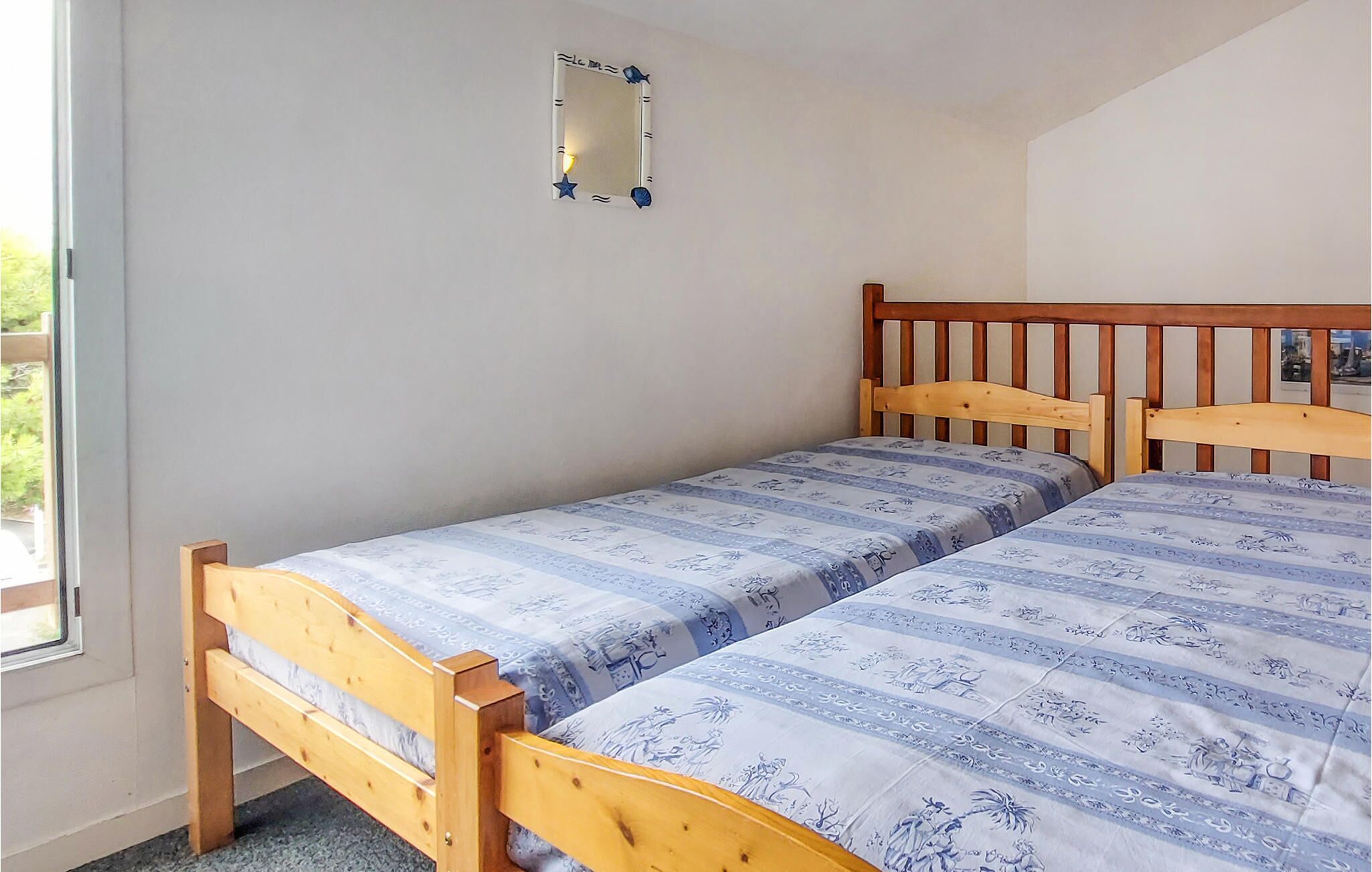 A small, single-bed room with a wooden headboard, a small mirror on the wall, and a window providing natural light.