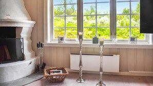 Ein gemütliches Zimmer mit Kamin, einem großen Fenster mit Blick ins Grüne und zwei Kerzenständern auf dem Fensterbrett.