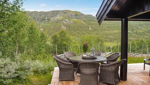 Eine Holzterrasse mit Korbmöbeln und einem Glastisch, mit Blick auf eine gebirgige Landschaft.