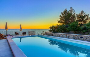Una piscina con vista sul mare al tramonto, circondata da lettini e ombrelloni.