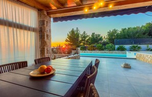 An outdoor patio with a table set for two, a stone wall, and a view of a swimming pool and trees. - 3 bedroom stunning home in Ivan Dolac (Ivan Dolac)