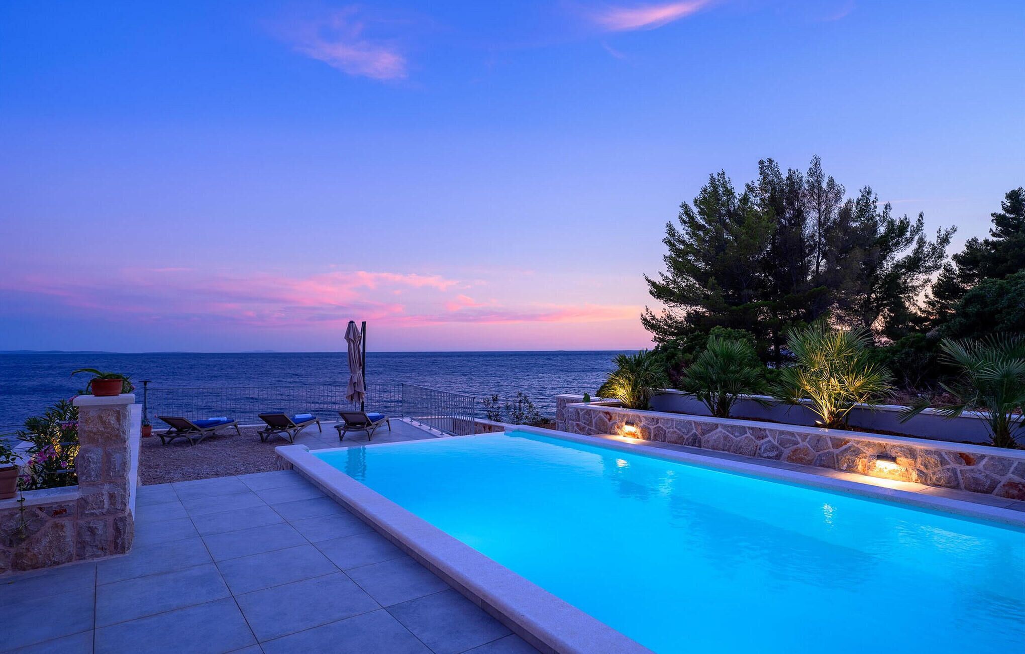 A pool area with lounge chairs, a stone wall, and a view of the sea at dusk.