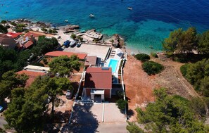 Una vista aerea di una proprietà costiera con piscina, vialetto in pietra e un litorale roccioso con barche.