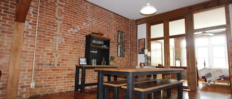 A room with exposed brick walls, wooden beams, a large table with benches, and a hanging light fixture.