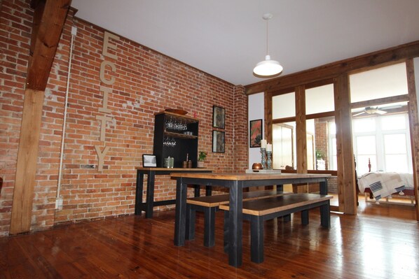 A room with exposed brick walls, wooden beams, a large table with benches, and a hanging light fixture.