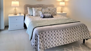 Una cama bien tendida con ropa de cama estampada, dos lámparas de noche, un tapiz de macramé y una mesita de noche blanca pequeña con cajones.
