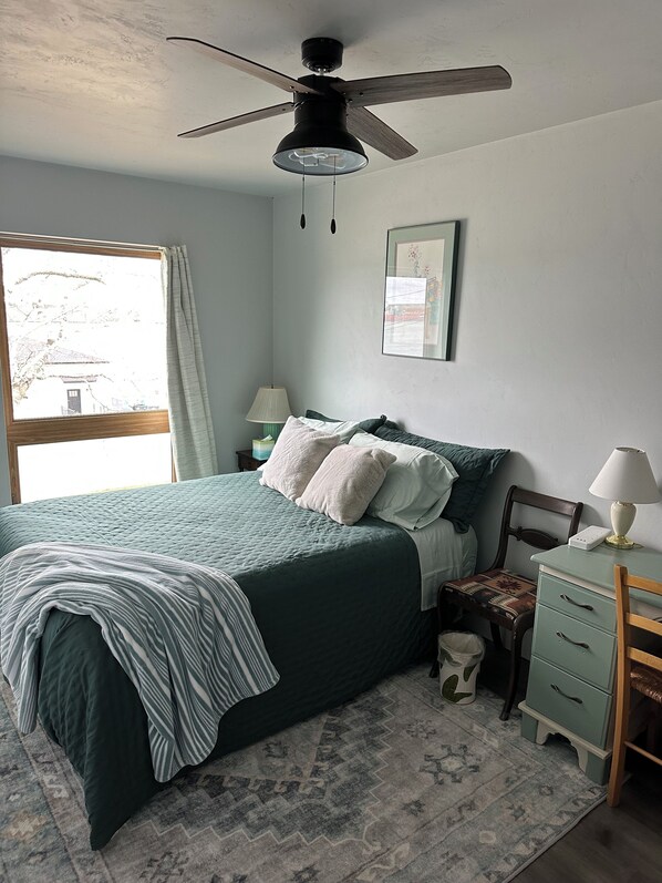 A bedroom with a bed, a desk, a chair, a lamp, a ceiling fan, a window, and a framed picture on the wall. - Water views from every window, covered porch & private deck, 7 min walk to beach (Sturgeon Bay)