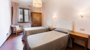 Une chambre d’hôtel avec deux lits, une armoire en bois, une chaise en osier, une fenêtre avec des rideaux et un luminaire suspendu.