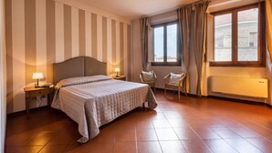 Habitación de hotel con cama, dos mesitas de noche con lámparas, una silla, una ventana con cortinas y un radiador.