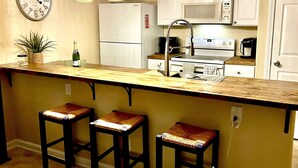 Una cocina con una isla central, taburetes de bar, microondas, refrigerador e iluminación colgante.