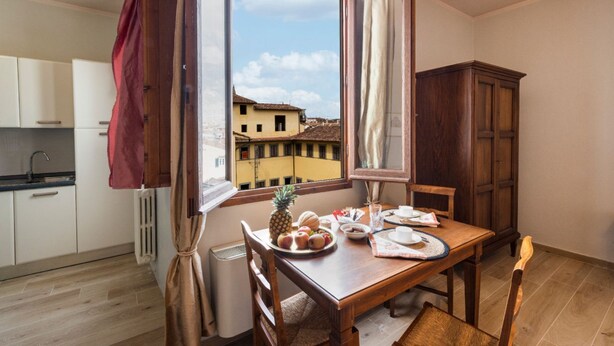 Un coin repas avec une table dressée pour deux personnes, une fenêtre avec des rideaux, une vue sur des immeubles et une cuisine en arrière-plan.