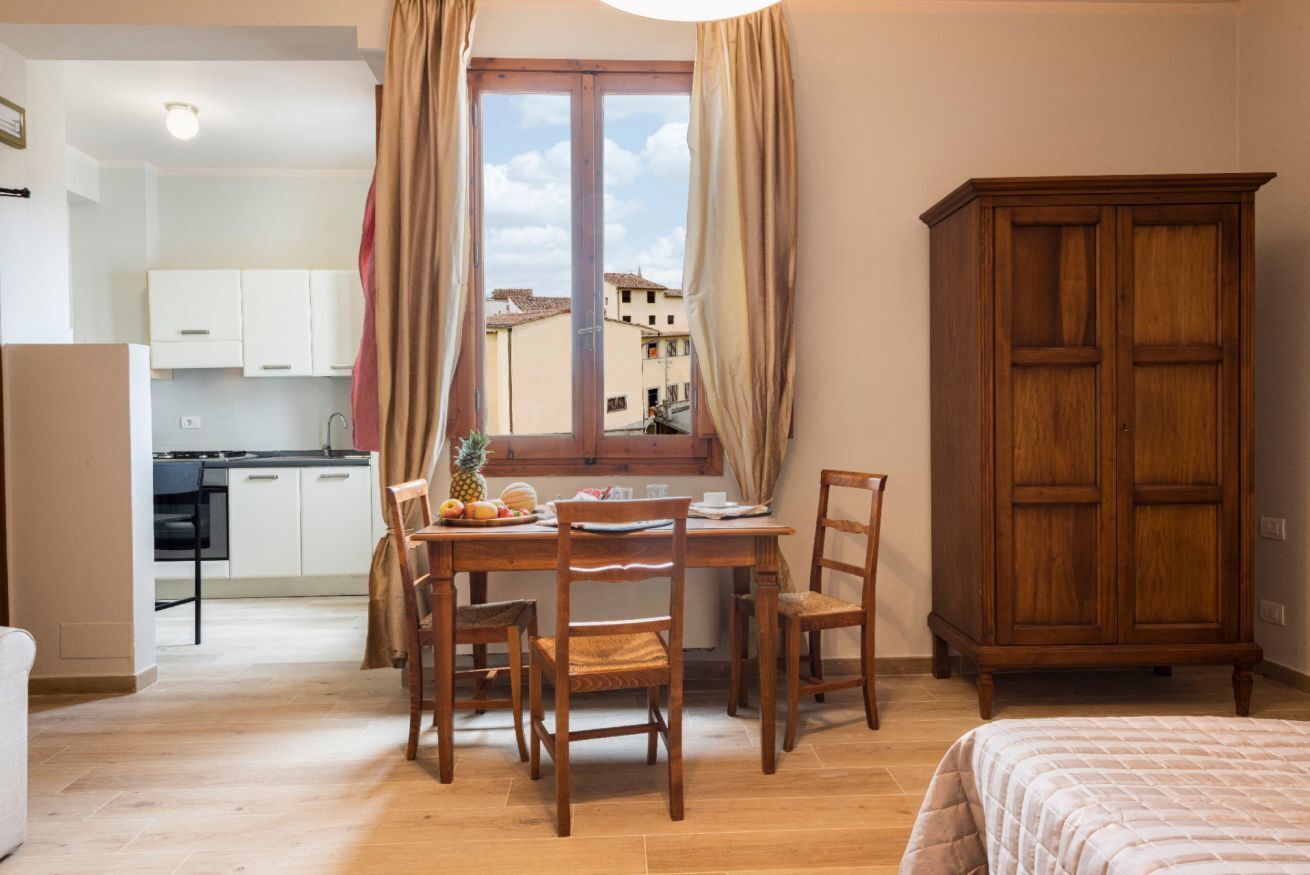 A room with a dining table, chairs, a window with curtains, a wooden cabinet, and a kitchenette in the background.