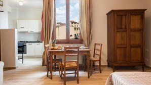 Une pièce comprenant une table à manger, des chaises, une fenêtre avec des rideaux, un buffet en bois et, au fond, une petite cuisine.
