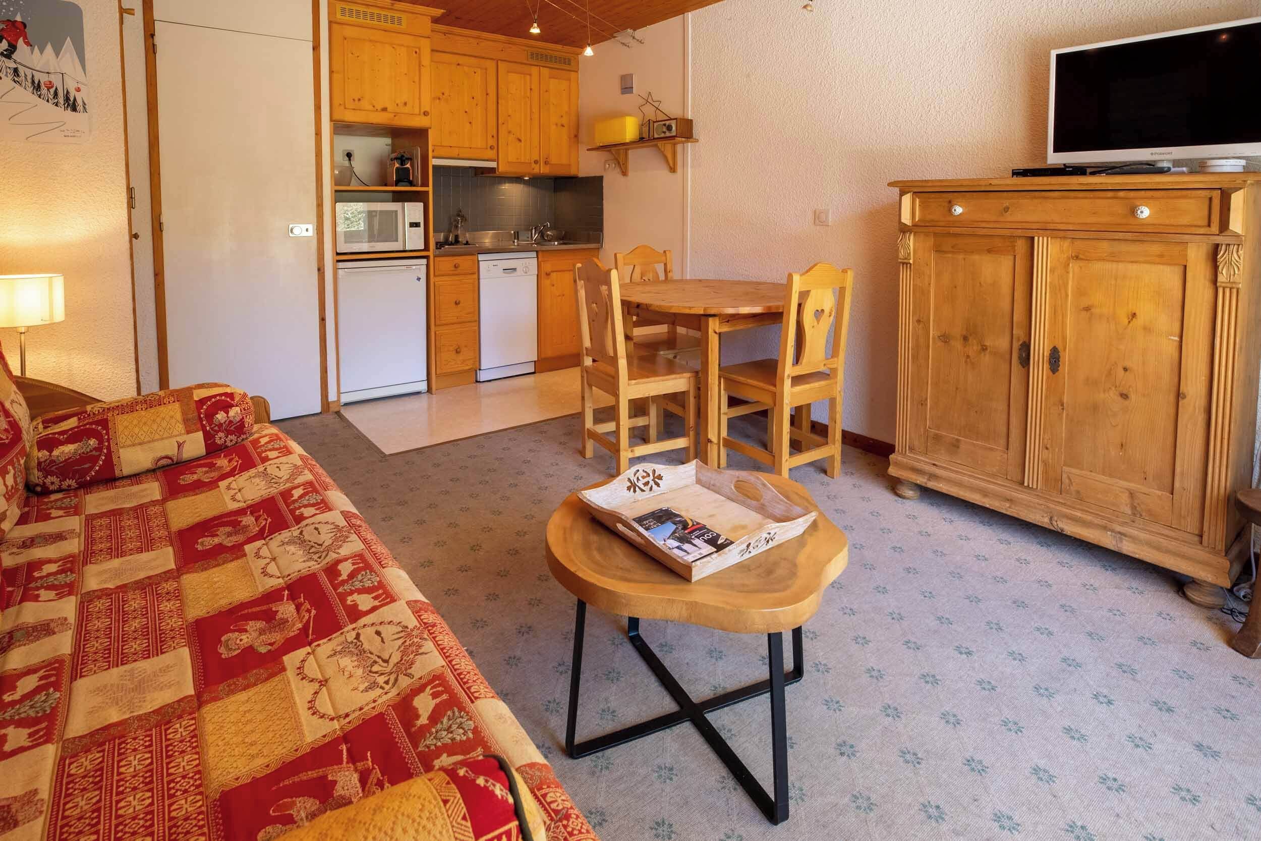 Ett mysigt rum med en soffa i trä, ett litet träbord med ett tidningsställ, ett matbord i trä med stolar, en köksdel med träskåp, en inbyggd TV och ett träskåp med lådor.