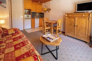 Ein gemütliches Zimmer mit einer Holzsofa, einem kleinen Holztisch mit einer Zeitschrift, einem Holzessttisch mit Stühlen, einer Küchenzeile mit Holzschränken, einem eingebauten Fernseher und einer Kommode mit Schubladen.