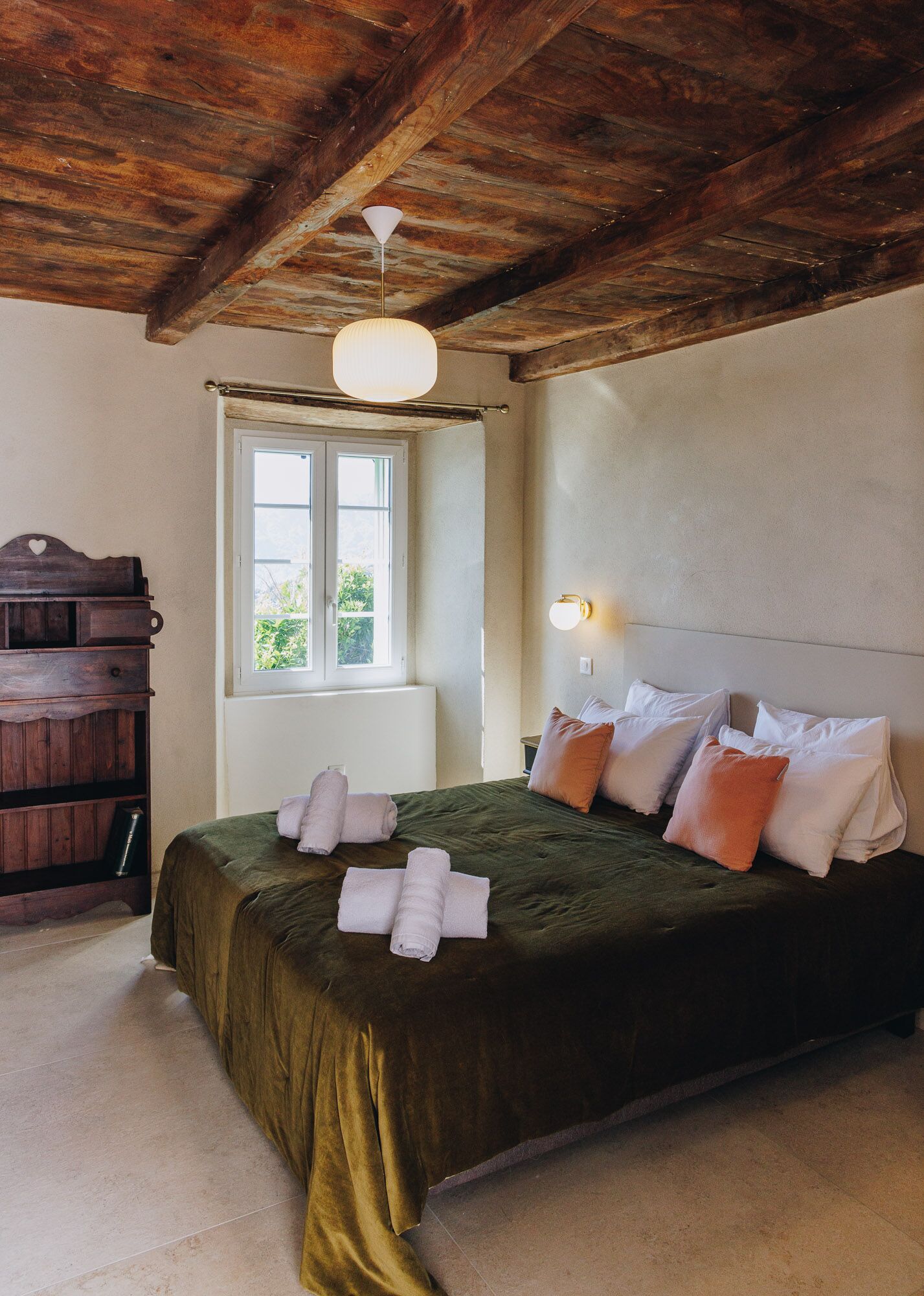 Un dormitorio con techo de madera, una cama con ropa de cama verde y múltiples almohadas, una mesita de noche, una ventana con vistas y un aparador de madera.