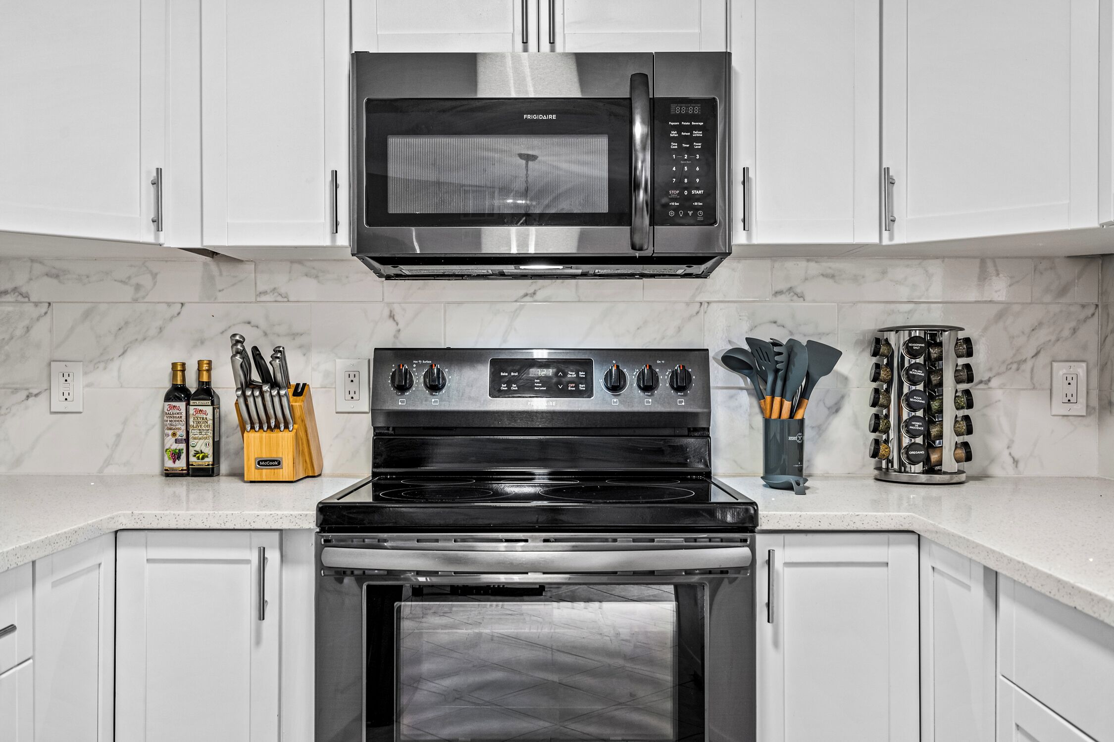 A kitchen with white cabinets, a stainless steel microwave, a stove, and a built-in oven. There are bottles of olive oil, a knife block, and a spice rack on the counter.