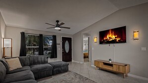 Una sala de estar con un sofá modular, un televisor de pantalla plana sobre un mueble de madera, un ventilador de techo y una chimenea.