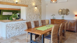A dining area with a wooden table and wicker chairs, a stone wall, and a kitchen in the background. - Awesome home in Violes (Violes)
