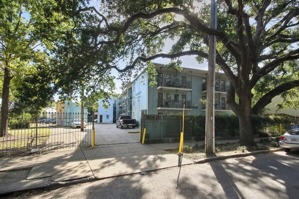 A residential building with a gated entrance, a parking area, and a large tree in the foreground.