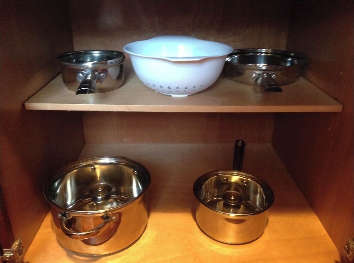 A kitchen shelf with stainless steel pots and a white colander.