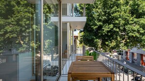 Ein moderner Balkon mit Holztisch und -stühlen, Glaswänden und Blick auf eine von Bäumen gesäumte Straße.