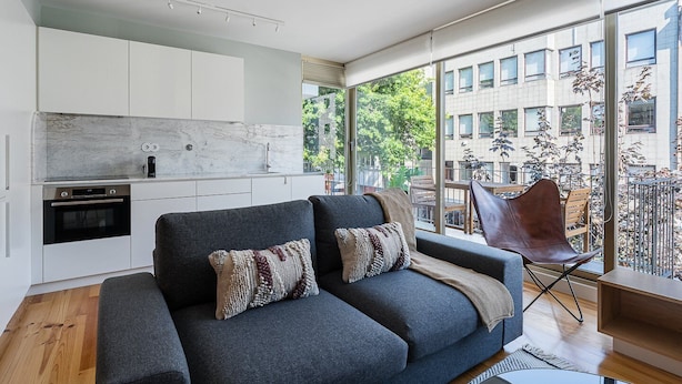 Ein modernes Wohnzimmer mit grauem Sofa, einer Kaffeemaschine und Blick auf einen Balkon mit Pflanzen sowie ein Gebäude im Hintergrund.