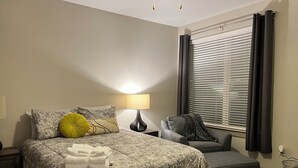 A hotel room with a bed, a nightstand, a lamp, a grey sofa, and a window with blinds. - 5840 #312 Cozy but Accessible Hideaway. Lots of Amenities (Chapel Hill)