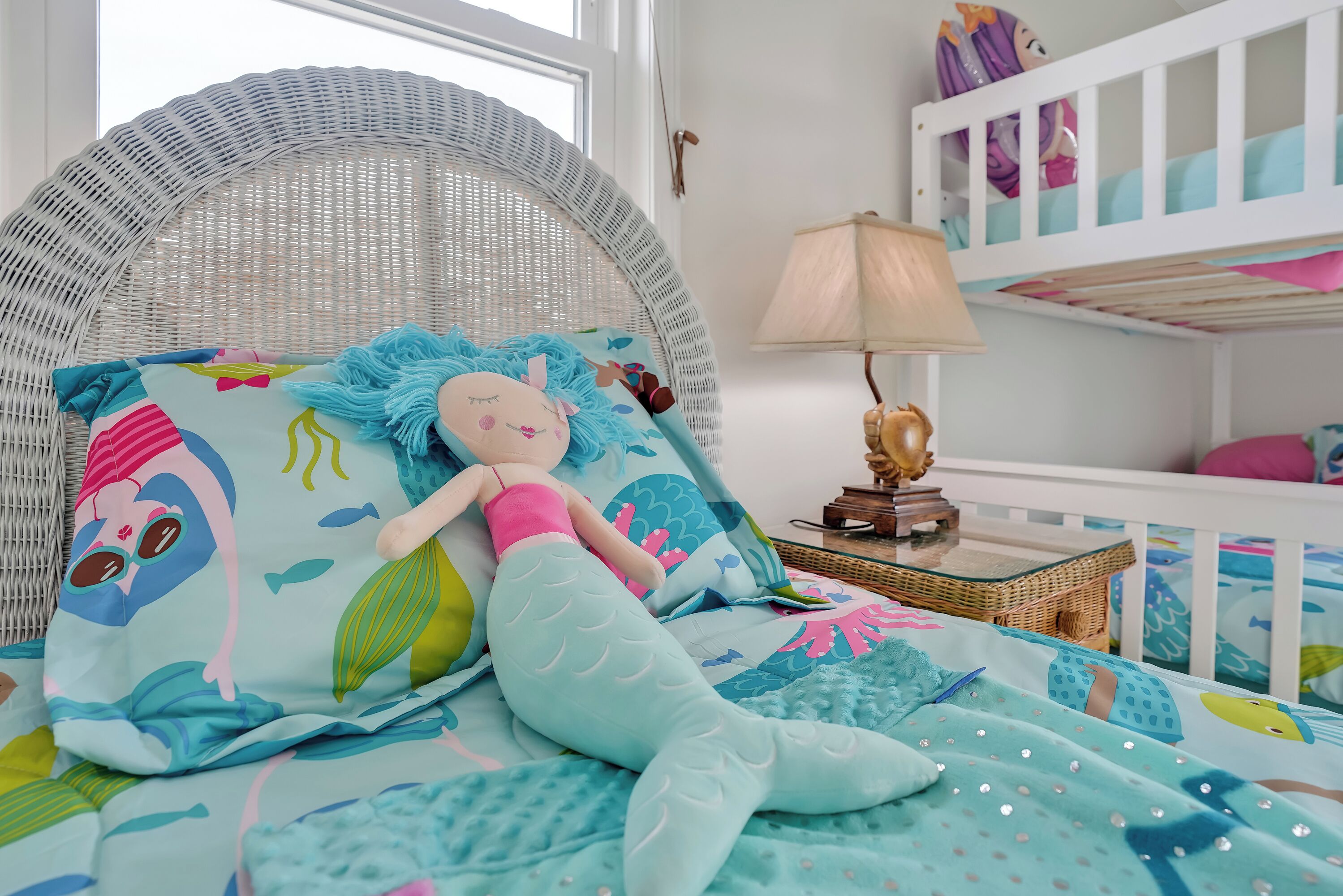 Un lit d’enfant avec une literie sur le thème de la mer, une chaise en osier, une lampe et, au fond, un lit superposé.