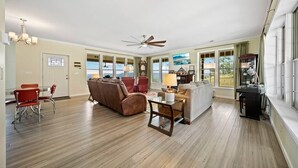 A spacious living room with a wooden floor, a ceiling fan, a dining area with red chairs, and a kitchen area with a black countertop.