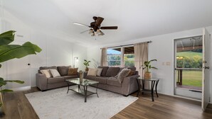 Ein Wohnzimmer mit Ecksofa, einem Glastisch und einem Deckenventilator. Ein großes Fenster mit Vorhängen bietet Blick auf Grünflächen.