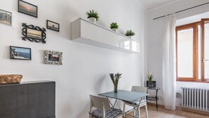 A dining area with a table, chairs, a small side table with a plant, and a white shelf above with potted plants.