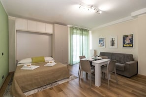 Um quarto com cama, escrivaninha, cadeira, sofá cinza e mesa de jantar.