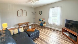 Ein Wohnzimmer mit einem schwarzen Ledersofa, einem Holztisch, einem Fernseher, einer Stehlampe und einem Fenster mit Vorhängen.
