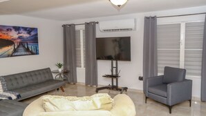 A living room with a grey sofa, a cream-colored armchair, a flat-screen TV, and a painting of a sunset over a pier. - EcoMar Rentals #1: Cabo Rojo, Puerto Rico; Beach & Pool (Cabo Rojo)
