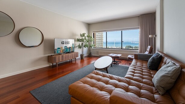 Una sala de estar moderna con un sofá de cuero, una mesa de centro y un televisor sobre un mueble de madera. Hay un ventanal con vistas al océano y al paisaje urbano.