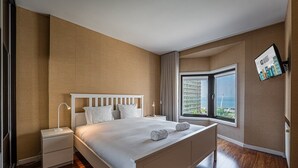 Una habitación de hotel moderna con una cama grande, una mesita de noche, una lámpara de noche y un ventanal con vistas a un edificio y al mar.