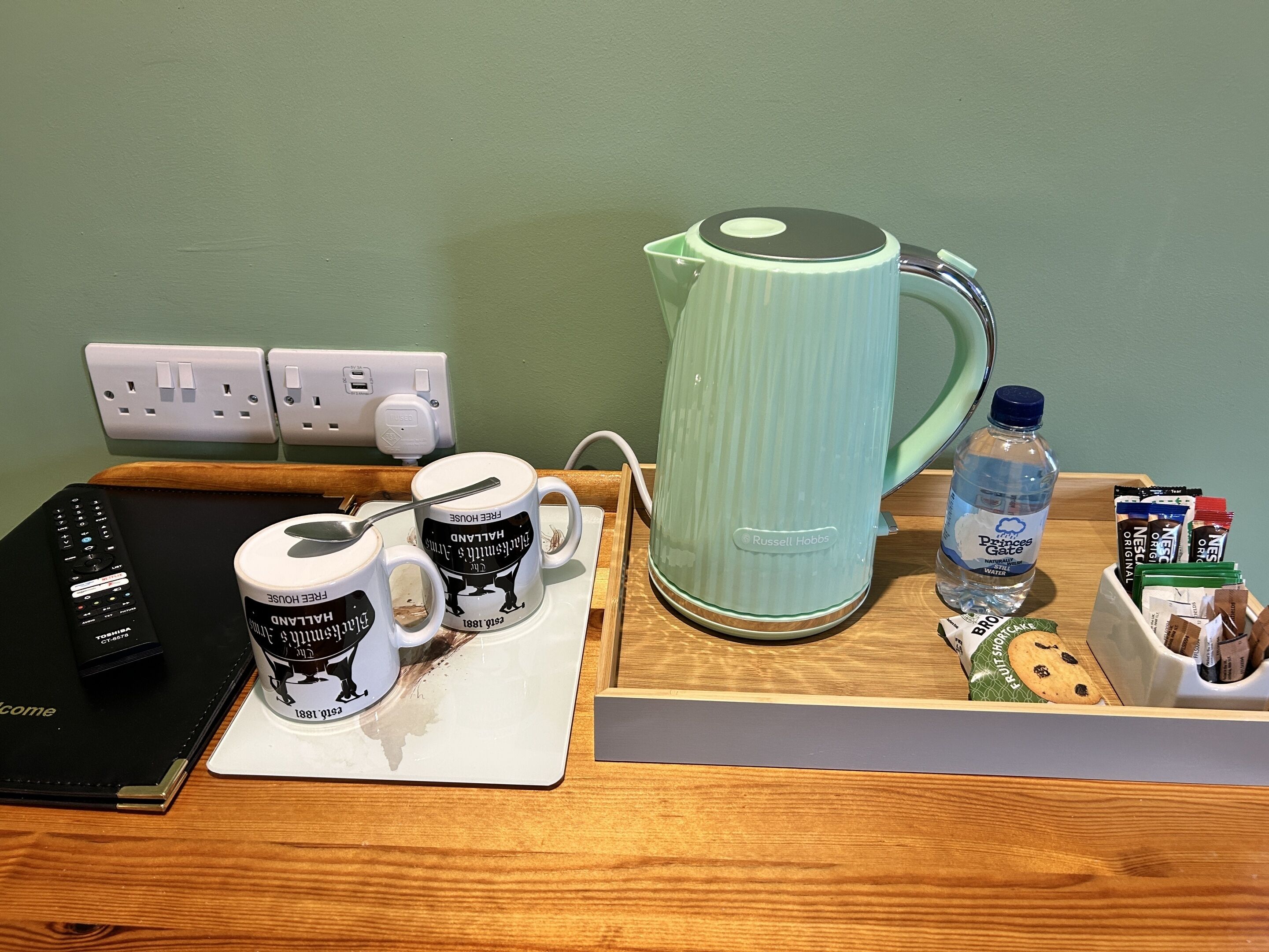 Una encimera de cocina con una tetera, tazas de café, un control remoto, una botella de agua y una caja de bolsitas de té.