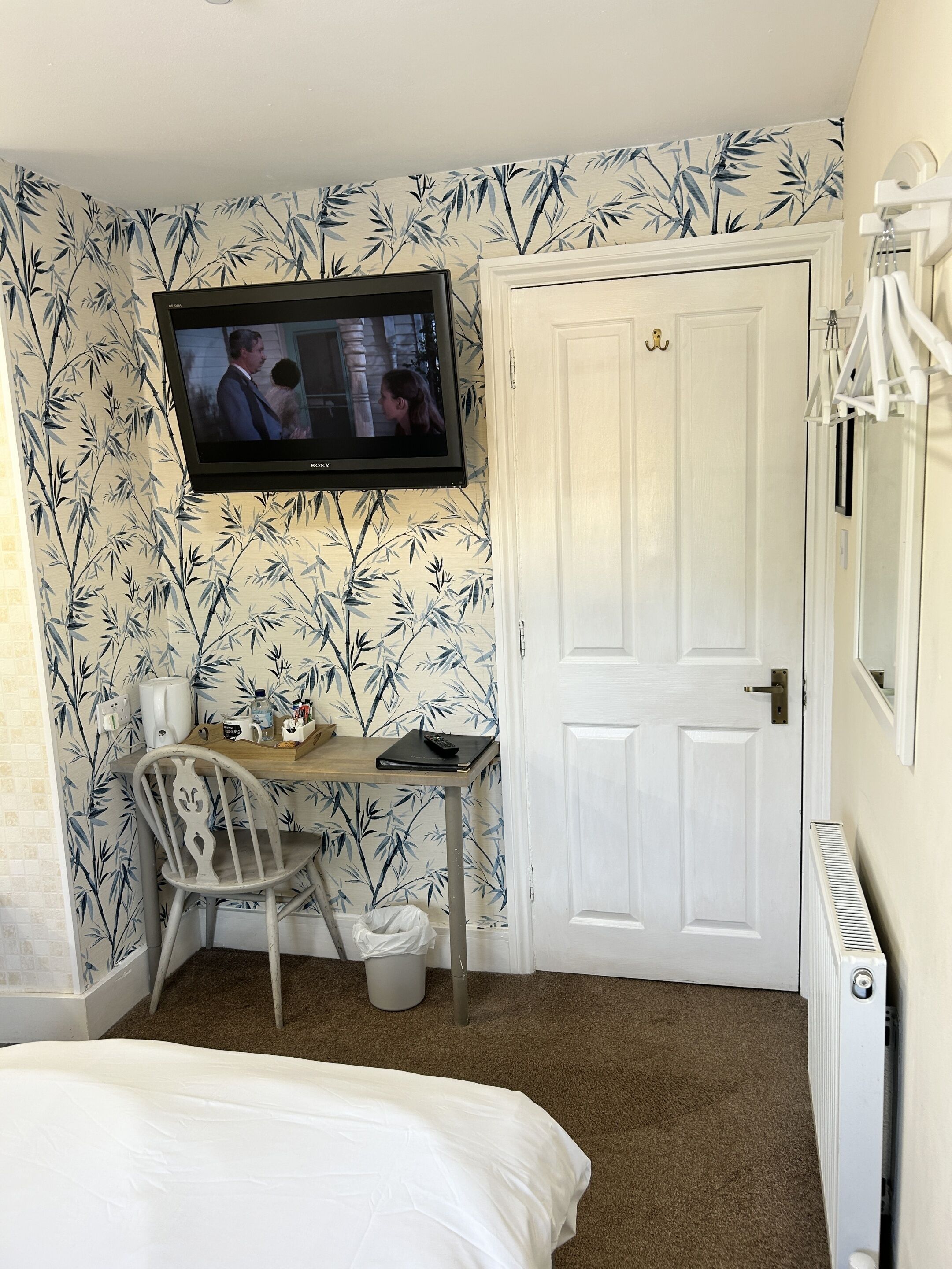 A small, neatly arranged bedroom with a bed, a desk with a chair, a television mounted on the wall, and a door.