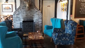 A cozy room with a brick fireplace, two blue armchairs, a plaid coffee table, and a stack of firewood.