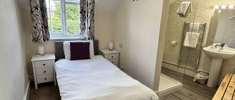 A bedroom with a bed, a nightstand, a window with curtains, a bathroom with a sink and mirror, and a shower area.