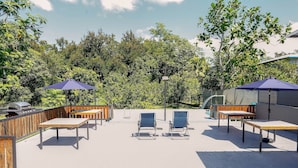 Une terrasse extérieure avec des tables et des chaises en bois, deux parasols bleus et un barbecue.