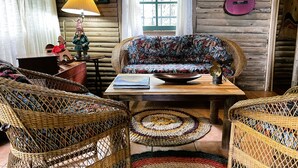 Una habitación acogedora con una pared de troncos de madera, un sofá tejido y colorido, sillas de mimbre, una pequeña mesa con un libro, una lámpara y una guitarra.