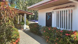 A residential building with a covered porch, a door with a decorative emblem, and a pathway lined with flowering plants. - Villa 1 minute walking to beach with pool A/C pet friendly (Jaco)
