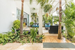 A modern indoor garden with tropical plants, a wooden deck, and a seating area with a black sofa and blue cushions.