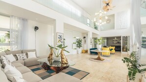 A spacious living area with a sectional sofa, a coffee table, and a dining table. There are plants, yellow chairs, and a ceiling fan.